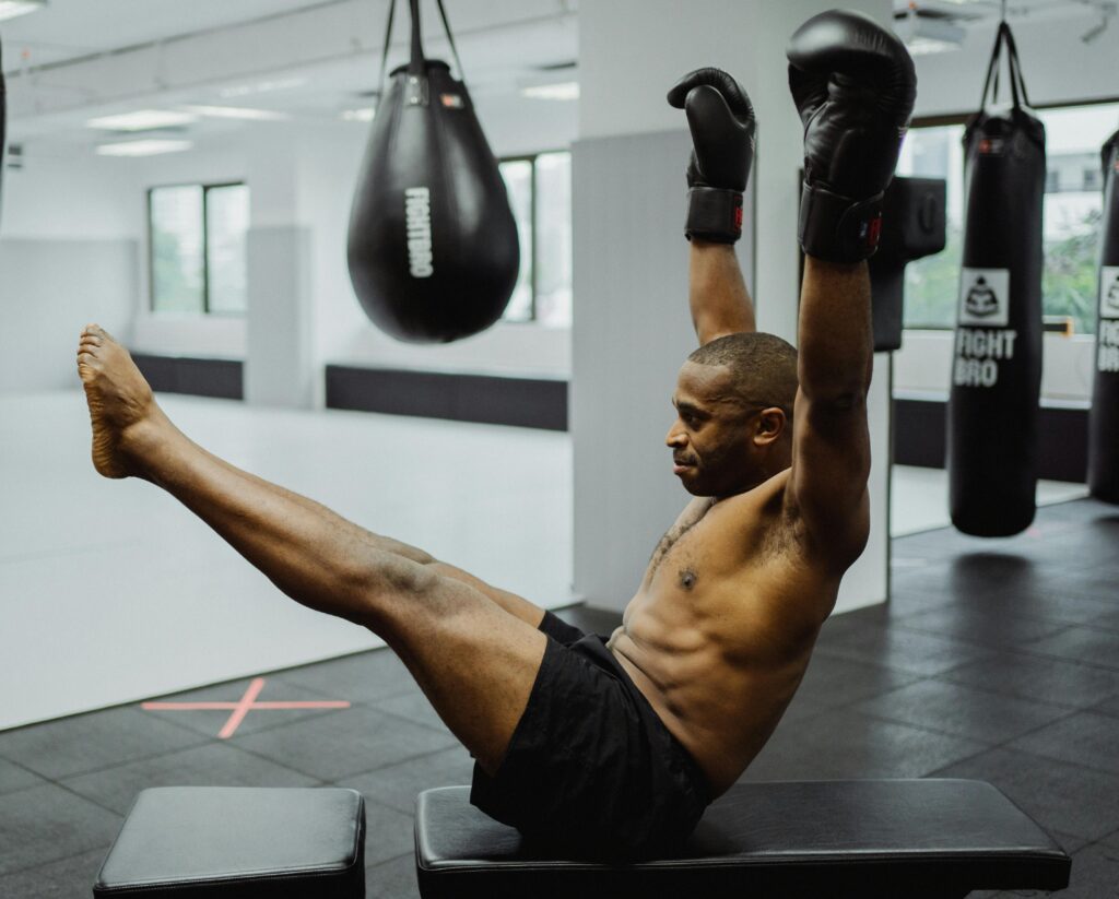 Shirtless man in boxing gloves stretching legs during workout in gym with punching bags.