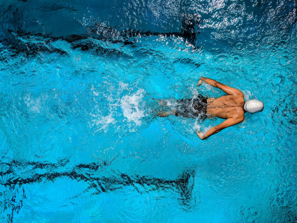 Top view of a swimmer wearing a cap, performing a front crawl stroke in a clear blue swimming pool.